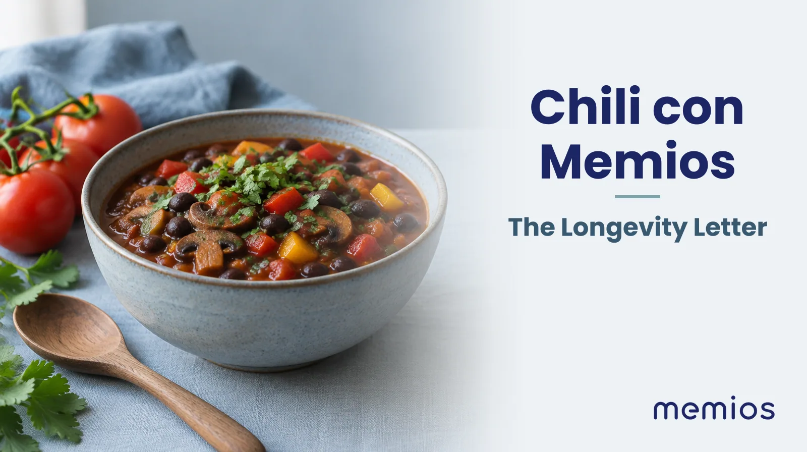 Photograph for Chili con Memios: a ceramic bowl of chili with black beans, peppers and mushrooms, with a wooden spoon and a few fresh tomatoes beside it.